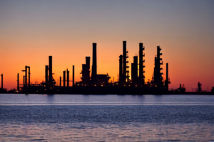 Oil refinery silhouette at sunset over water, symbolizing oil price volatility amid geopolitical tensions